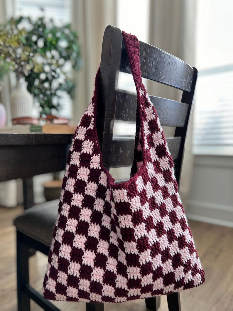 Peut inclure: Un sac fourre-tout crochet&eacute; &agrave; la main avec un motif en damier bordeaux et rose clair. Le sac est suspendu &agrave; une chaise en bois fonc&eacute;. La bandouli&egrave;re et le bord sup&eacute;rieur du sac sont bordeaux.