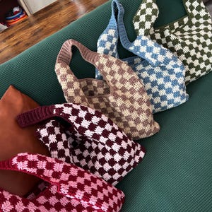 Peut inclure: Plusieurs sacs fourre-tout crochet&eacute;s &agrave; la main, aux couleurs vari&eacute;es : marron, bleu, vert et rouge. Chaque sac pr&eacute;sente un motif &agrave; carreaux et une longue bandouli&egrave;re. Les sacs sont dispos&eacute;s sur une surface verte.