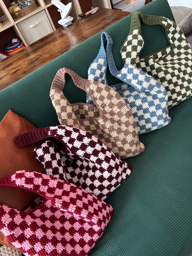 Peut inclure: Plusieurs sacs fourre-tout crochet&eacute;s &agrave; la main, aux couleurs vari&eacute;es : marron, bleu, vert et rouge. Chaque sac pr&eacute;sente un motif &agrave; carreaux et une longue bandouli&egrave;re. Les sacs sont dispos&eacute;s sur une surface verte.
