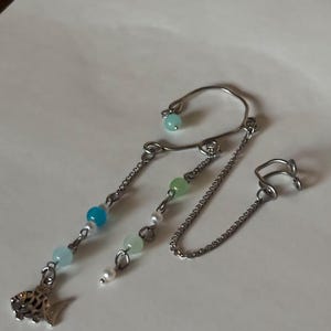 May include: A silver ear cuff with a chain and dangling beads in shades of blue, green, and white. A small fish charm hangs from the bottom. The ear cuff has a curved design that wraps around the ear.
