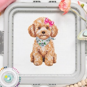 May include: An embroidery hoop containing white fabric with a machine-embroidered design of a light brown curly-haired puppy. The puppy wears a white bandana with green leaves and a small blue flower, and a bright pink bow on its head.
