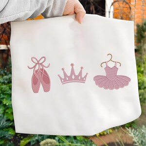 May include: White fabric with embroidered pink ballet-themed designs. The designs include a pair of ballet shoes, a crown, and a tutu on a hanger. The embroidery is detailed and the overall aesthetic is delicate.
