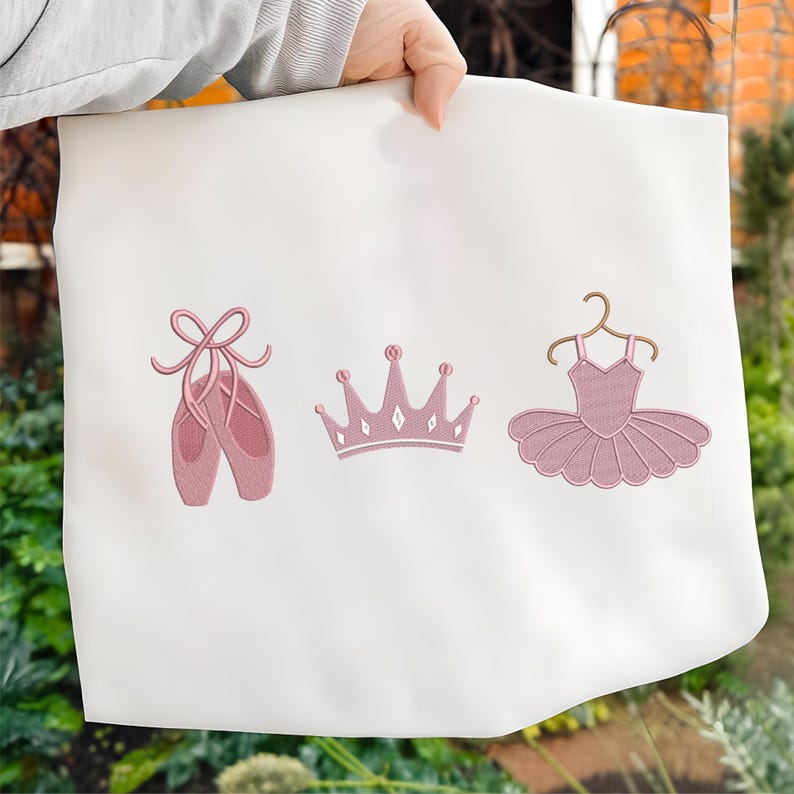May include: White fabric with embroidered pink ballet-themed designs. The designs include a pair of ballet shoes, a crown, and a tutu on a hanger. The embroidery is detailed and the overall aesthetic is delicate.