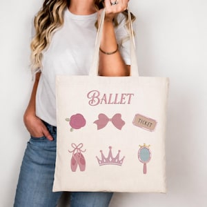 May include: A beige tote bag featuring the word "Ballet" in pink script. The bag is decorated with pink ballet-themed illustrations, including a rose, bow, ticket, pointe shoes, crown, and hand mirror. The bag is held by a person.