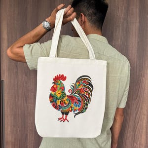 May include: Cream-colored tote bag featuring a colorful rooster design. The rooster is depicted with vibrant red, yellow, blue, and green details. The bag has two white handles and is held by a person.