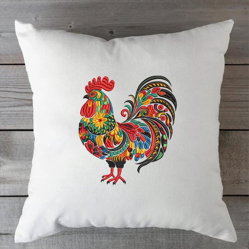 May include: White square pillow featuring a colorful embroidered rooster design. The rooster has a red comb, yellow and green body, and a vibrant tail with red, blue, and black details. The pillow is on a wooden surface.