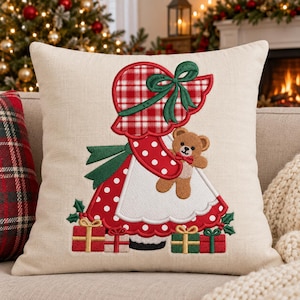 May include: A beige square throw pillow featuring an embroidered design of a figure wearing a red and white plaid bonnet and a red dress with white polka dots. The figure holds a brown teddy bear, surrounded by small wrapped gift boxes.