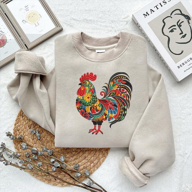 May include: Beige sweatshirt featuring a vibrant embroidered rooster design. The rooster has a red comb, and a colorful tail with shades of yellow, green, blue, and red. The sweatshirt has a ribbed crew neck and cuffs.