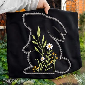 May include: Black tote bag with a bunny silhouette and embroidered floral details. The design includes white daisies, yellow accents, and green leaves, outlined with a white lace trim. The text "EMBRICHMOND" is at the bottom.