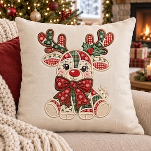Puede incluir: Un cojín cuadrado de tela beige claro con un diseño de reno bordado. El reno presenta astas y cuerpo estilo patchwork con patrones en rojo, verde y blanco, un gran lazo rojo y una pequeña rama de acebo con bayas rojas.