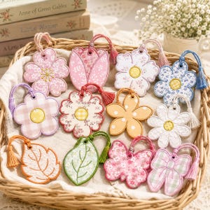 May include: A collection of twelve decorative hanging ornaments in a woven basket, featuring various floral, leaf, and butterfly shapes. Each ornament is crafted with fabric patterns, embroidered details, and a matching coloured hanging tassel.