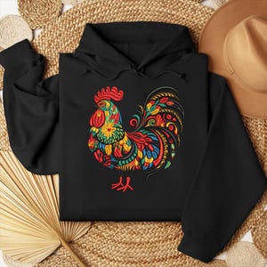 May include: Black hooded sweatshirt featuring a colorful rooster design. The rooster is adorned with red, yellow, green, and blue floral and patterned details. The design is centered on the front of the sweatshirt.