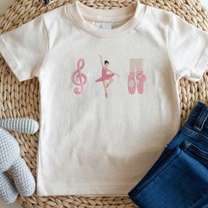 May include: Cream-colored t-shirt featuring a pink treble clef, a ballerina in a pink tutu, and a pair of pink ballet shoes. The shirt is paired with blue denim jeans. The design is likely for a child.