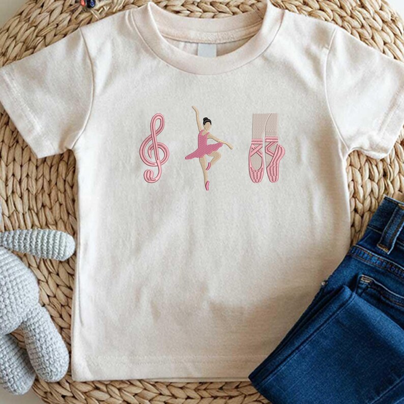 May include: Cream-colored t-shirt featuring a pink treble clef, a ballerina in a pink tutu, and a pair of pink ballet shoes. The shirt is paired with blue denim jeans. The design is likely for a child.