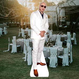 May include: A life-size cardboard cutout of a man in a white suit and sunglasses stands on a grassy area. The man is wearing a white double-breasted suit, a navy blue shirt, and brown shoes. The background shows tables set for an outdoor event.