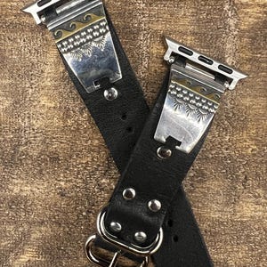 May include: A black leather watch band featuring silver-tone metal connectors and decorative plates. The plates are adorned with engraved wave patterns and sunburst motifs in silver and gold tones. The band includes a silver-tone metal buckle.