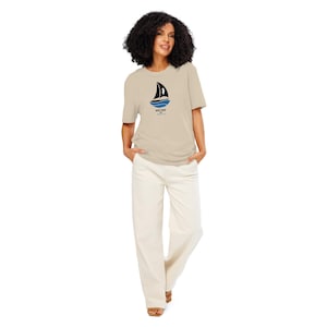 Puede incluir: Una camiseta beige con un gráfico de velero negro y el texto "White Sail" en negro. La modelo lleva la camiseta con pantalones blancos.