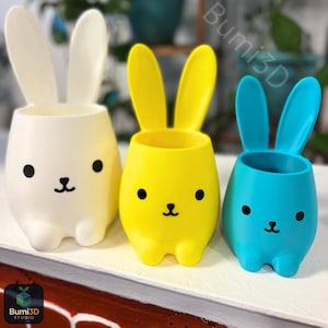 May include: Three colorful bunny-shaped planters. The planters are in white, yellow, and blue. Each planter has large ears, a round body, and a cute bunny face with black eyes and a small black nose. The planters are made of plastic.