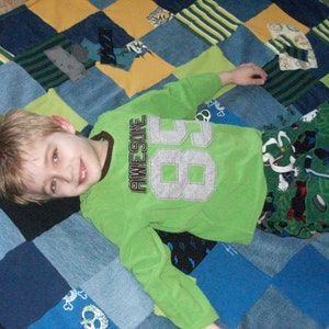 May include: A young person wearing a green shirt with the word "AWESOME" and a large number "86" on it, lying on a patchwork quilt made of blue denim and green fabric.