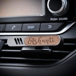 May include: A car air vent diffuser with a wooden central section engraved with the name "Ashanti" in a cursive font. The diffuser has a silver metal frame with three ventilation slots. It is installed in a car's dashboard.