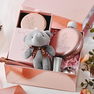 以下が含まれることがあります： 茶色のリボンが付いた小さな灰色のテディベア、コンパクトミラー、フラスコ、ペン、小さな金属製のカップが入った、ローズゴールドのギフトボックス。ボックスはピンクの紙で満たされ、花の模様で飾られています。