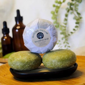 May include: A close-up of three green shampoo bars. One is wrapped in white paper with a label that reads "Matcha + Tallow Shampoo Bar." Two unwrapped bars sit on a black soap dish. Amber glass bottles are in the background.