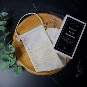 May include: A natural cotton soap organizer with a wooden handle and a mesh pocket, presented on a wooden tray. The packaging reads "COTTON SOAP ORGANIZER". The background is a dark, marble-like surface.