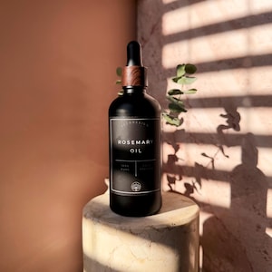 Puede incluir: Una botella de vidrio negro de aceite de romero con tapa y cuentagotas de madera. La etiqueta dice "Rosemary Oil" y "100% Pure". La botella est&aacute; sobre un pedestal de piedra, con luz solar y sombras en el fondo.