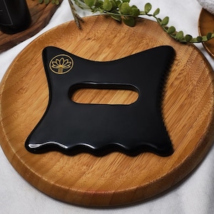 Puede incluir: Una herramienta de masaje gua sha negra con un logotipo de flor de loto dorada, sobre un plato de madera. La herramienta tiene una forma curva con un recorte de mango y un borde ondulado. El plato de madera es redondo y tiene vetas naturales.