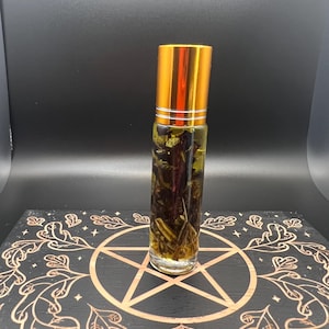 Puede incluir: Un frasco roll-on de vidrio transparente con tapa dorada, lleno de un líquido amarillo y elementos botánicos. El frasco está sobre una superficie de madera negra con un pentagrama dorado y diseño de hojas.