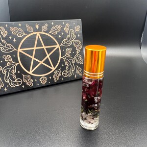 Könnte beinhalten: Eine transparente Glas-Rollerflasche für ätherische Öle mit goldenem Deckel, gefüllt mit getrockneten Blumen und Kristallen. Eine schwarze Holztafel mit einem goldenen Pentagramm und Blattdesign befindet sich im Hintergrund.
