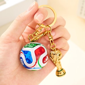 Puede incluir: Un llavero dorado con un balón de fútbol y un trofeo. El balón es blanco con detalles rojos, azules y verdes. El trofeo es una réplica detallada. El llavero está en la mano de una persona.