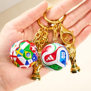 Puede incluir: Dos llaveros de balón de fútbol con cadenas y cierres dorados. Un balón presenta banderas de varias naciones, el otro tiene detalles en rojo, blanco y verde. Cada llavero incluye un pequeño trofeo dorado.