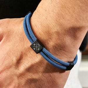 May include: A blue rope bracelet with a black square clasp. The clasp features white symbols. The bracelet is worn on a wrist. The bracelet is a stylish accessory.