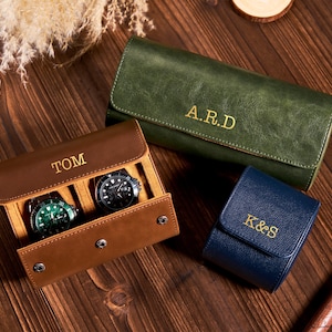 May include: Three leather watch cases in brown, green, and blue, each with gold lettering. The brown case is open, displaying two watches. The green case has the initials "A.R.D.", the blue case has "K&S", and the brown case has "TOM".