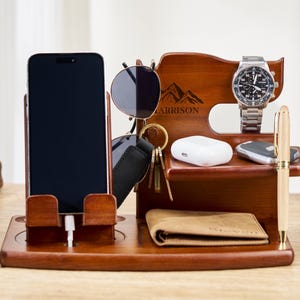 Puede incluir: Un organizador de escritorio de madera en un rico acabado marrón. Cuenta con un soporte para teléfono, un soporte para gafas de sol, una pantalla de reloj, un llavero y una pequeña cartera. También se ven un bolígrafo y unos auriculares inalámbricos.