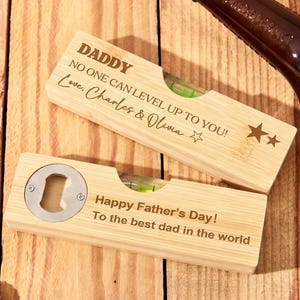 Könnte beinhalten: Zwei Holzflaschenöffner mit Wasserwaagen. Einer trägt die Aufschrift "DADDY" und "NO ONE CAN LEVEL UP TO YOU! Love, Charles & Olivia". Der andere sagt "Happy Father's Day! To the best dad in the world".
