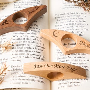 May include: Wooden book page holders with engraved designs and text. One is dark wood with "Kevin" and a mountain, another light wood with a moon and stars, and the last with "Just One More Page".