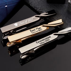 Può includere: Tre fermacravatte in metallo argentato e dorato. Uno recita "Class of 2025" con un'icona di cappello di laurea. Un altro recita "Best Man Devin 2025" e il terzo "Groomsman Ethan 2025".