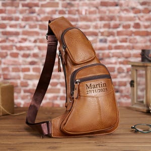 Könnte beinhalten: Eine braune Leder-Umhängetasche mit mehreren Reißverschlussfächern und einem verstellbaren Riemen. Auf der Tasche sind der Name "Martin" und das Datum "27/11/2025" eingraviert. Die Tasche liegt auf einer Holzoberfläche.