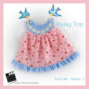 Peut inclure: Une robe bébé sans manches rose et bleue avec une bordure de marguerites et un ourlet à volants. La robe est sur un cintre avec des oiseaux bleus sur une ficelle au-dessus. Le texte "Daisy Top" et "Sizes NB - Toddler 3" est également visible.