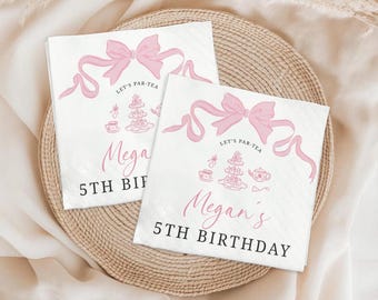 May include: Two white paper napkins with pink bow and ribbon designs. The napkins have the text "Let's Par-Tea", "Megan's", and "5th Birthday" printed on them. The napkins are on a woven, circular placemat.