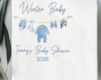 May include: A white square napkin with the text "Winter Baby" and "Tommy's Baby Shower 2026" in blue script. A clothesline illustration features baby clothes, including a hat, mittens, and a onesie.