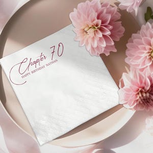 May include: A white paper napkin with a diamond pattern, featuring the text "Chapter 70 HAPPY BIRTHDAY NATHAN" in burgundy script. The napkin is placed on a light brown plate, adorned with pink dahlias.