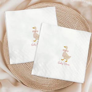 May include: Two white paper napkins with a diamond pattern. Each napkin features a watercolor illustration of a goose wearing a pink bow, with the word "Baby" and a name in pink script. The napkins are on a woven tray.
