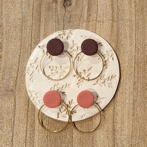 Puede incluir: Cuatro pares de pendientes de aro hechos a mano. Dos pares presentan tachuelas circulares de color marrón oscuro, y los otros dos tienen tachuelas de color coral. Los aros son de color dorado y se exhiben sobre una placa de cerámica blanca con motivos florales.