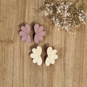 Puede incluir: Cuatro cortadores de galletas con forma de flor en lila claro y blanco roto. Están dispuestos sobre una superficie de madera, con una ramita de flores blancas en la esquina superior derecha. Los cortadores son de material sólido.