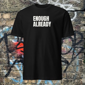 Op de afbeelding: Zwart t-shirt met de witte tekst "ENOUGH ALREADY" op de borst gedrukt. Het shirt is een korte mouw ronde hals stijl. De achtergrond is een bakstenen muur met graffiti.