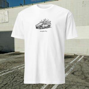 May include: White t-shirt featuring a black line drawing of a vehicle engulfed in flames. The text "dumpster fire" is printed below the graphic. The shirt has short sleeves and a crew neck.