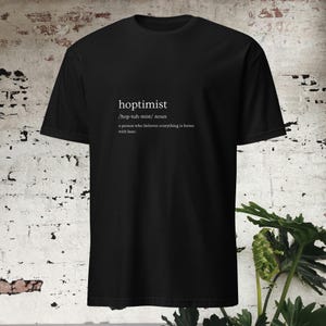 Op de afbeelding: Zwart T-shirt met witte tekst die "hoptimist /hop-tuh-mist/ noun a person who believes everything is better with beer." luidt.